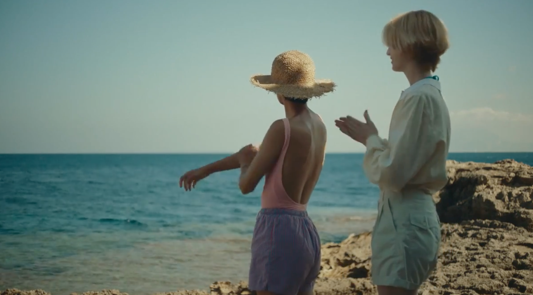 Two people stand on a rocky shore by the sea. One wears a straw hat, pink swimsuit, and purple shorts; the other claps and wears a light shirt and shorts. The weather appears sunny and clear.