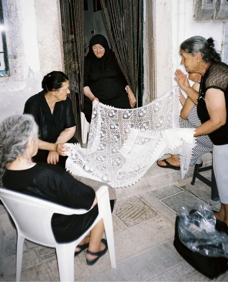 Four elderly women, dressed mostly in black, sit and stand outside a stone building, admiring a large, intricate white lace cloth together. One woman stands in a doorway behind the others.