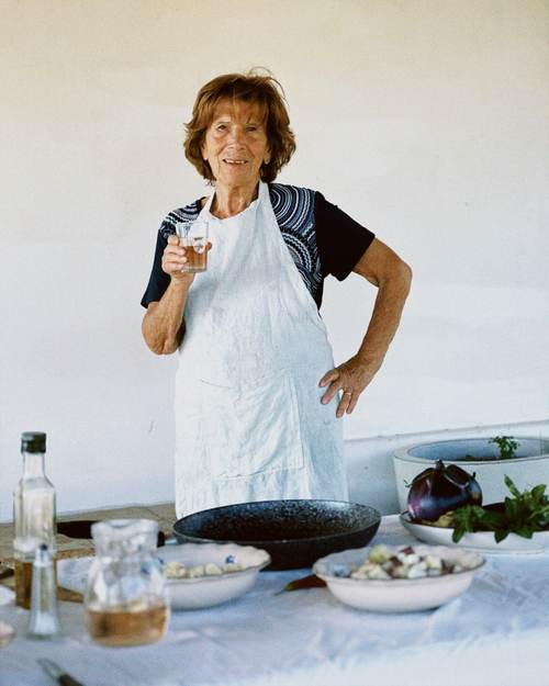 An older woman wearing a white apron stands at a table with various bowls, a bottle of olive oil, and fresh ingredients. She holds a glass and smiles at the camera, with a white wall in the background.