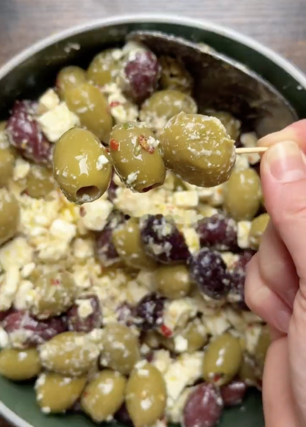 A hand holds a toothpick skewered with three green olives above a bowl of mixed green and black olives tossed with feta cheese.