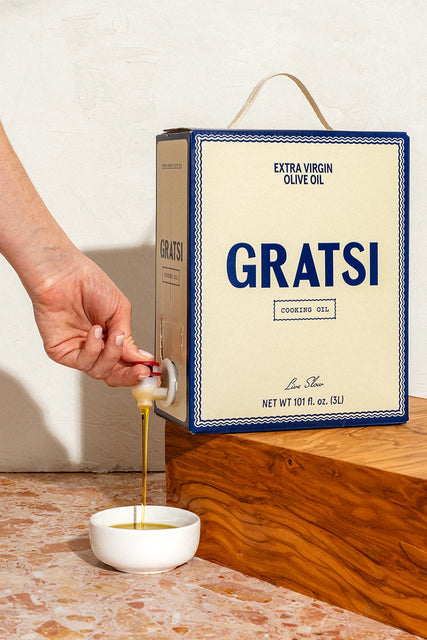 A hand turns the spigot on a boxed container of Gratsi Cooking Oil, 100% Greek Extra Virgin Olive Oil, pouring a stream into a small white bowl.