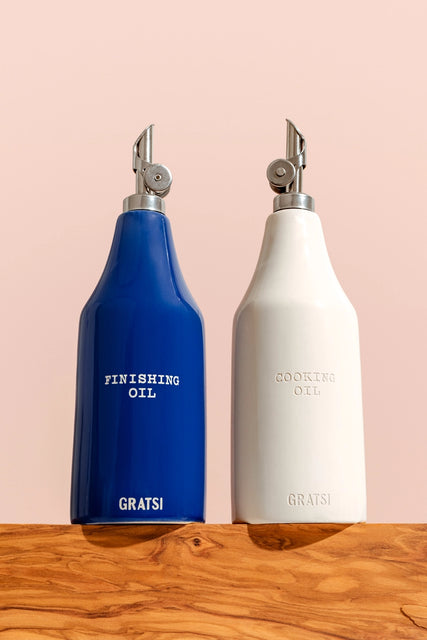 The Oil Cruet Set by Gratsi features two ceramic bottles with metal spouts: a blue Finishing Oil and a white Cooking Oil, both labeled Gratsi at the bottom, displayed on a wooden surface.