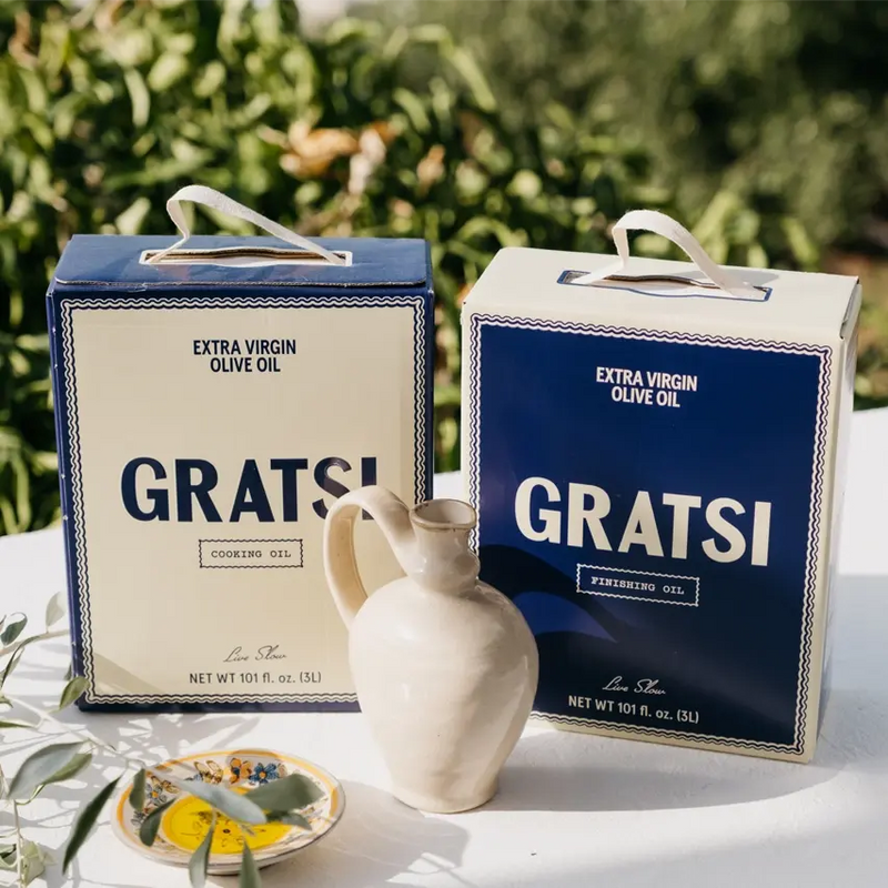 Two rectangular cartons of Gratsi extra virgin olive oil, one labeled Cooking Oil and the other Finishing Oil, sitting on a table next to a ceramic pitcher and a small floral saucer.