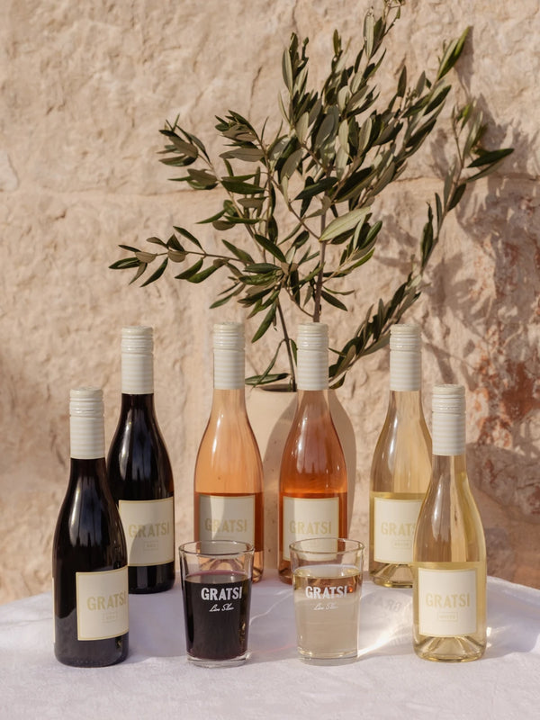 Six Gratsi wine bottles (red, rosé, and white) are displayed on a white tablecloth against a beige stone wall, with a leafy olive branch and three glasses—one with red wine, two with white—arranged in front.
