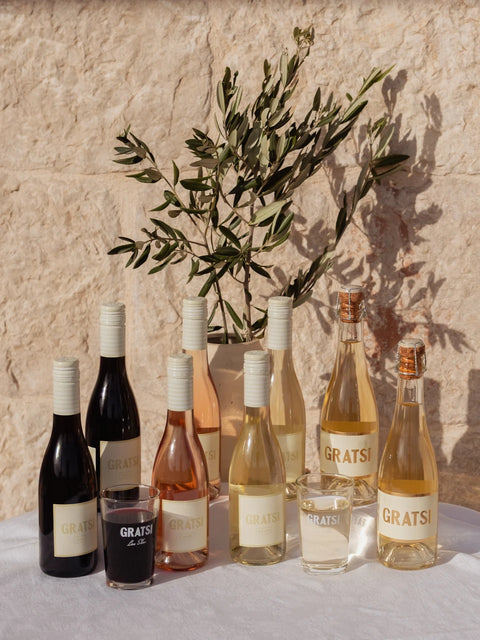 Eight bottles of GRATSI wine, two glasses (one with red wine, one with white), and a vase with olive branches are arranged on a white cloth against a sunlit stone wall.