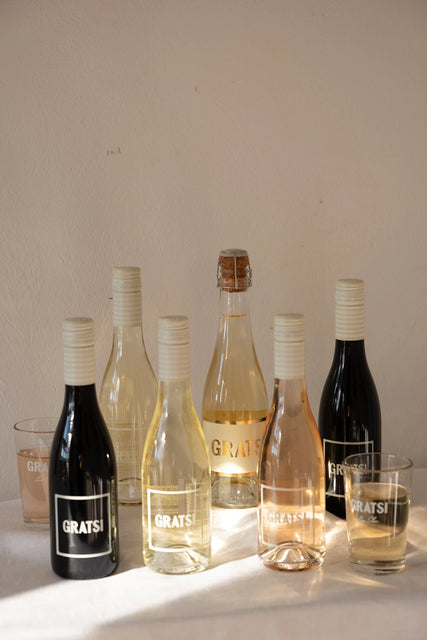 The Gratsi Wine Sampler Kit: Half Bottles + Glasses is displayed on a white surface, sunlight highlighting the variety of zero-sugar wines and the included branded glassware.