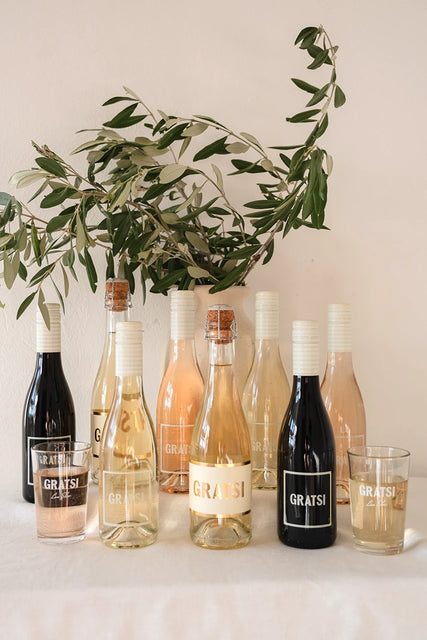 The Gratsi Wine Sampler Kit: Half Bottles + Glasses is displayed on a white surface, with two filled Gratsi glasses in front. Olive branches in a vase provide a decorative accent behind the bottles.