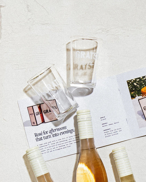 Three half wine bottles and two Gratsi Bàcaro glasses from the Wine Sampler Kit rest on a white surface next to an open booklet reading "Rosé for afternoons that turn into evenings," brightly lit in this online exclusive scene.