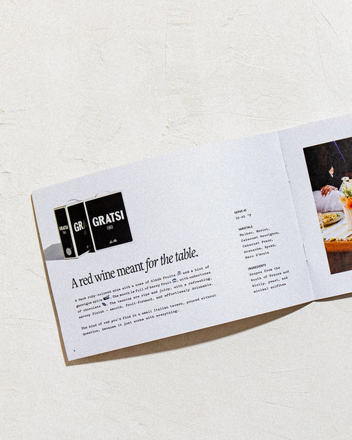 An open brochure on a textured white surface features Gratsi's Wine Sampler Kit: Half Bottles + Glasses, highlighting zero-sugar wines with text and wine box images on the left, and a partially visible dining table with Bàcaro glasses on the right.