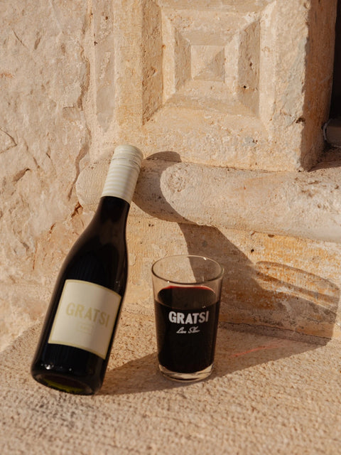 A bottle of GRATSI wine rests against a textured stone wall next to a glass filled with red wine, both illuminated by warm sunlight.