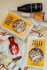 The Gratsi Wine & Pasta Valentine's Bundle features Zelli Pasta, a jar of Zelli pasta sauce, and Gratsi wine bottles, arranged on a cream surface with an orange-and-white striped cloth—perfect for gifting.
