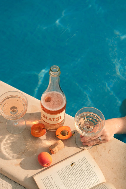 A bottle of Gratsi Sparkling Rosé Wine, two glasses, apricots, and an open book rest beside the pool. A hand lifts a glass of the delicate wine over the water as fine bubbles catch the sunlight. Hints of strawberry glide through the air while sunlight dances across the tranquil setting.