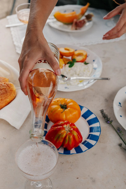 A hand pours Gratsi Sparkling Rosé Wine into a glass on a table arranged with heirloom tomatoes, sliced melon, cheese, fresh bread, and a sprig of lavender for a relaxed meal. Delicate fine bubbles and subtle strawberry notes from the Gratsi Sparkling Rosé add an elegant touch to the gathering.