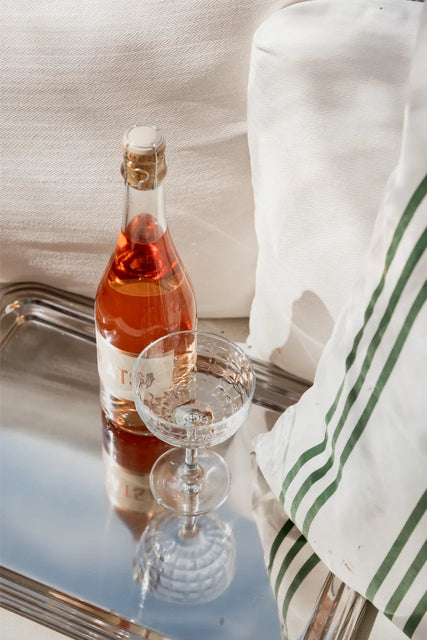 A bottle from the Gratsi Sparkling Variety Pack and an empty glass rest on a silver tray beside a white pillow with green stripes, all illuminated by gentle sunlight, evoking hints of delicate strawberry notes.