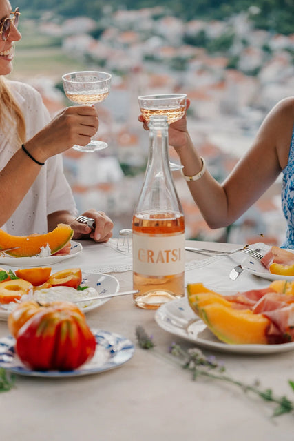 Two people toast with wine glasses at an outdoor table set with melon slices, tomatoes, and a bottle of Gratsi Sparkling Rosé Wine—celebrated for its fine bubbles and subtle strawberry notes—while a blurred townscape provides the backdrop.