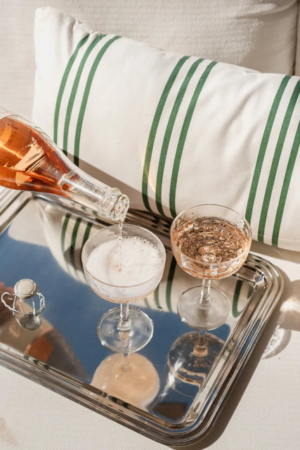A bottle from the Gratsi Sparkling Variety Pack is being poured into a coupe glass, next to another filled glass, both resting on a silver tray with a striped pillow in the background. The fine bubbles and delicate strawberry notes from Gratsi make this scene irresistibly elegant.