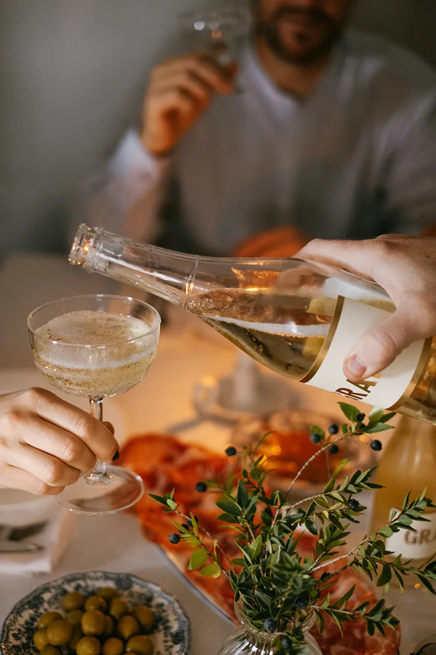 At a dinner table with olives, sliced meat, and greens, someone pours Gratsi Sparkling White Wine into a glass held by another guest while a third person sits in the background holding their own wine glass.