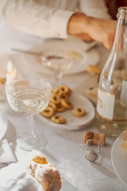 A table set with a glass of Gratsi Sparkling White, a bottle, snacks, a lit candle, and someone reaching for food—all bathed in warm, soft light.