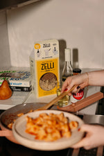 A person serves pasta with tomato sauce onto a plate in a kitchen. On the counter are a Gratsi Wine & Pasta Valentine's Bundle, a pear, some cookbooks, and a glass of white wine.