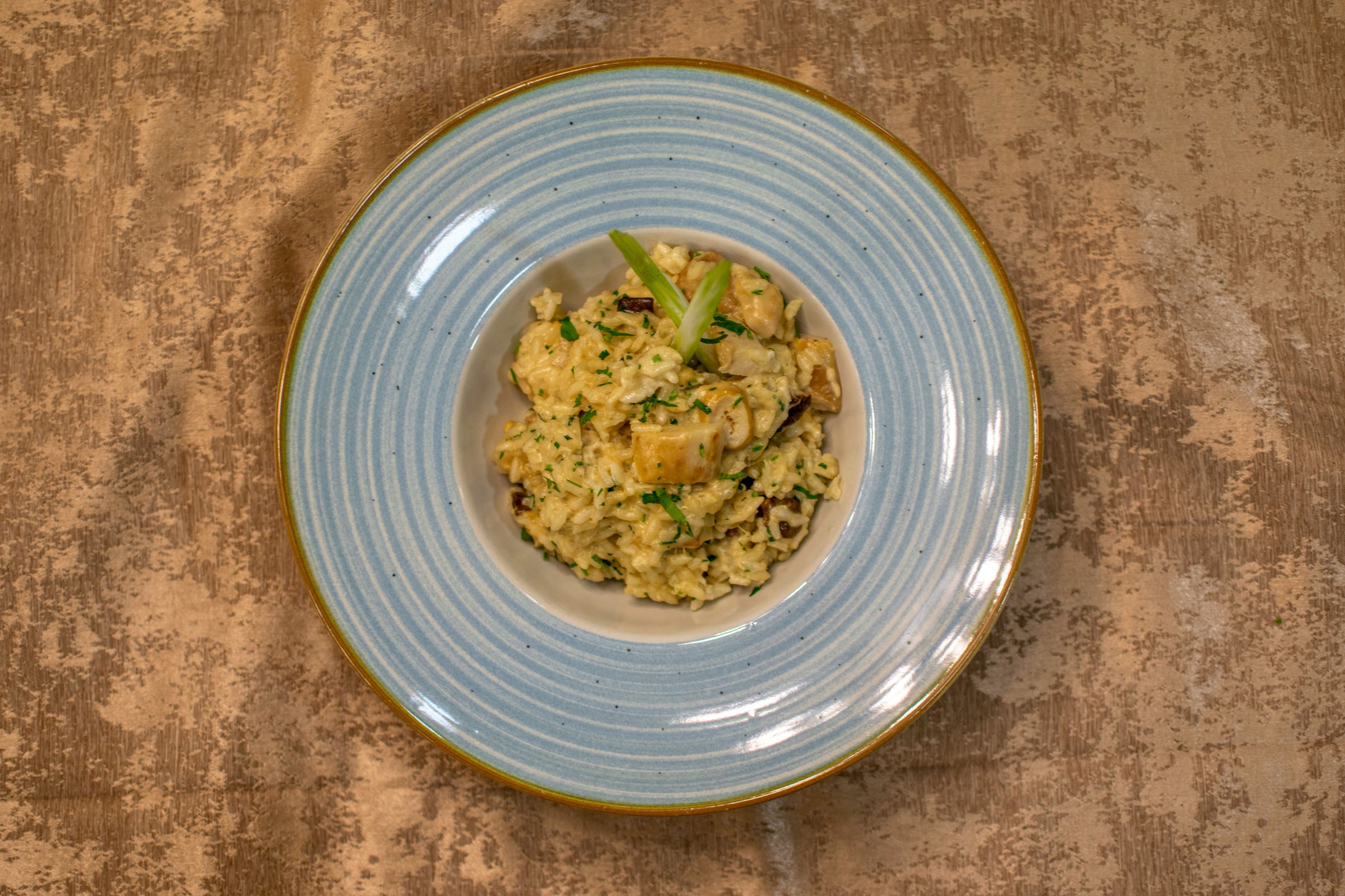 A plate of creamy risotto garnished with green herbs is served on a blue-rimmed dish, placed on a textured brown surface.