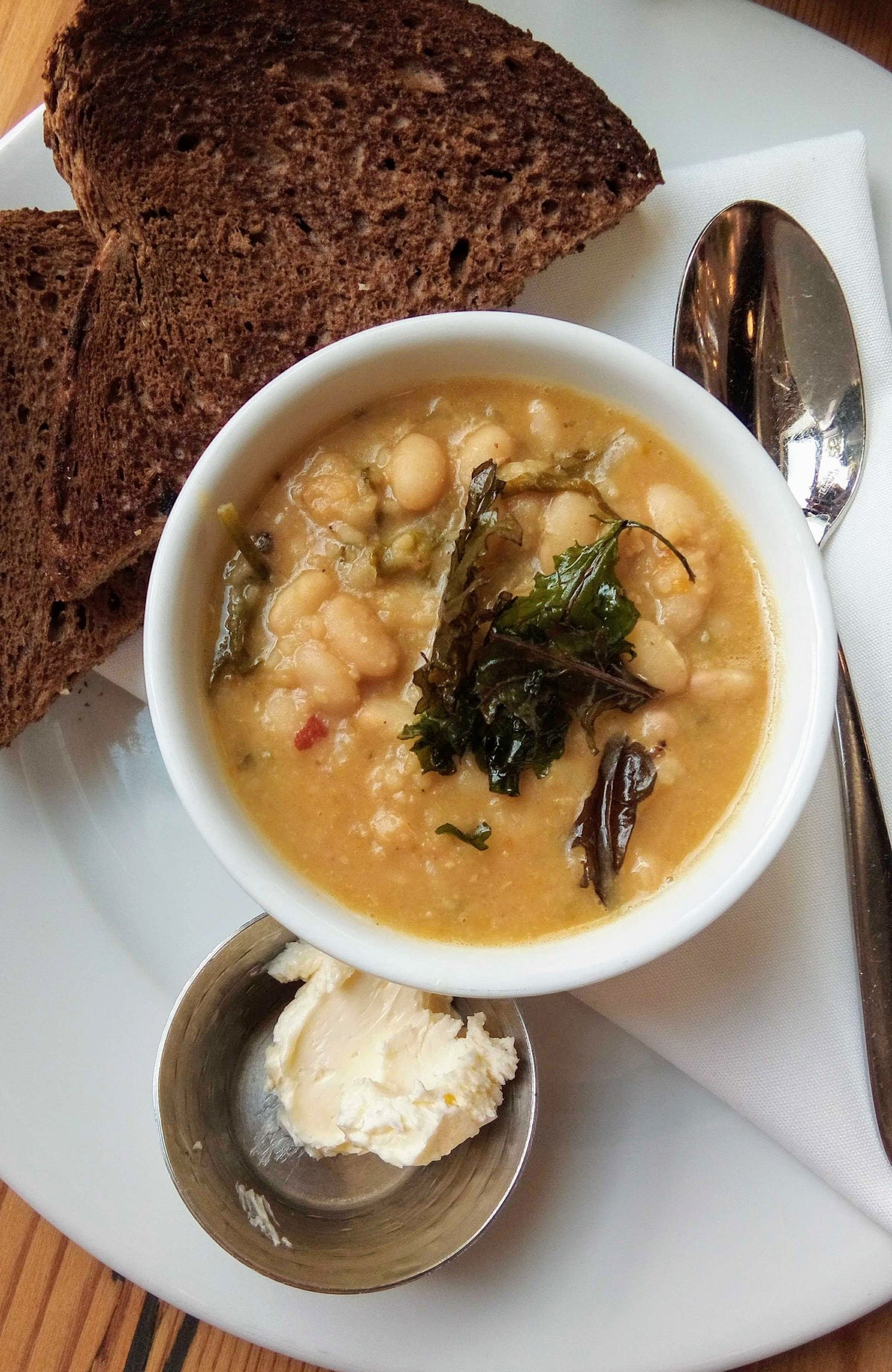 A bowl of bean soup garnished with greens, served with two slices of dark bread, a dollop of butter in a metal cup, and a spoon on a white plate.