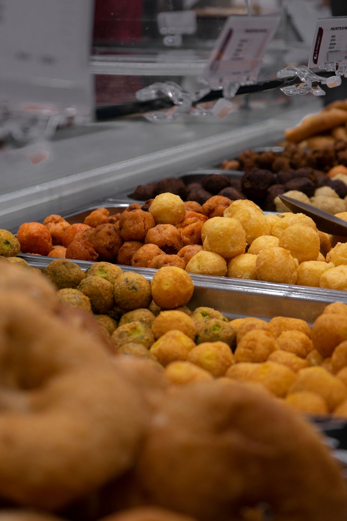 Assorted fried snacks and balls displayed in trays at a food counter, with different colors and textures visible. Some labels are seen above, slightly out of focus.