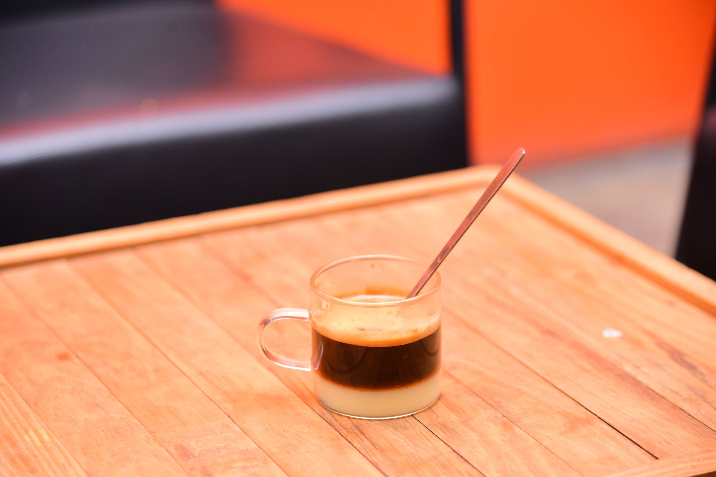 A clear glass cup of layered coffee with a metal spoon sits on a wooden table. The background is blurred with black seating and an orange wall visible.