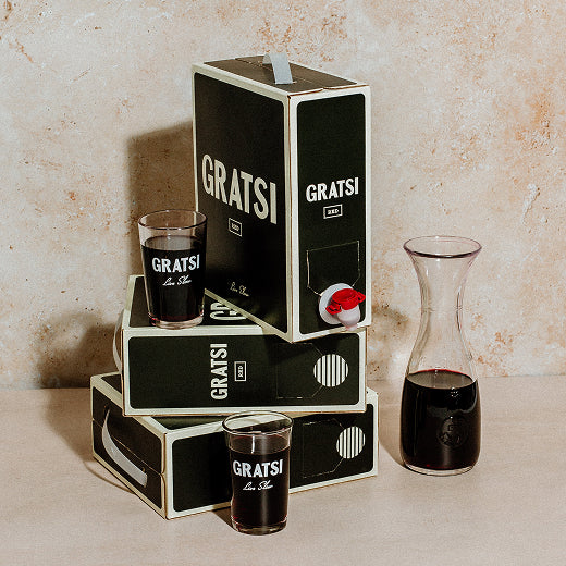 Stacked black boxes labeled GRATSI with a spout, two glasses filled with red wine, and a glass carafe with red wine, all set on a light textured surface.