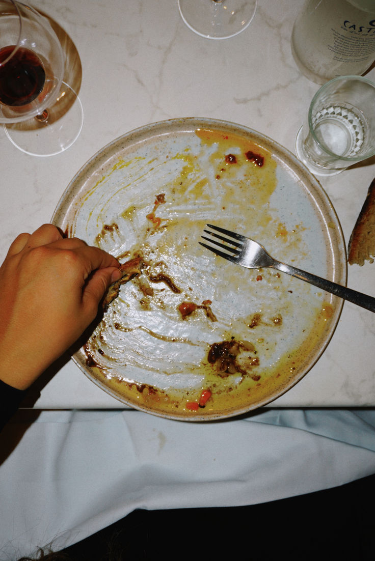 A hand wipes the last bits of sauce from an almost empty plate with bread, next to a fork, wine glasses, and a bottle on a marble table.