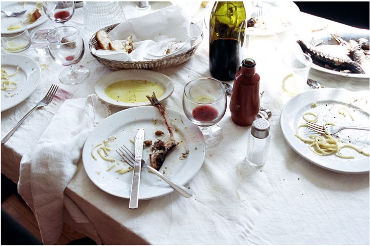 A dining table with messy, mostly empty plates, utensils, wine glasses with leftover red wine, a wine bottle, a bread basket, and scattered food remnants, indicating a meal has just been finished.