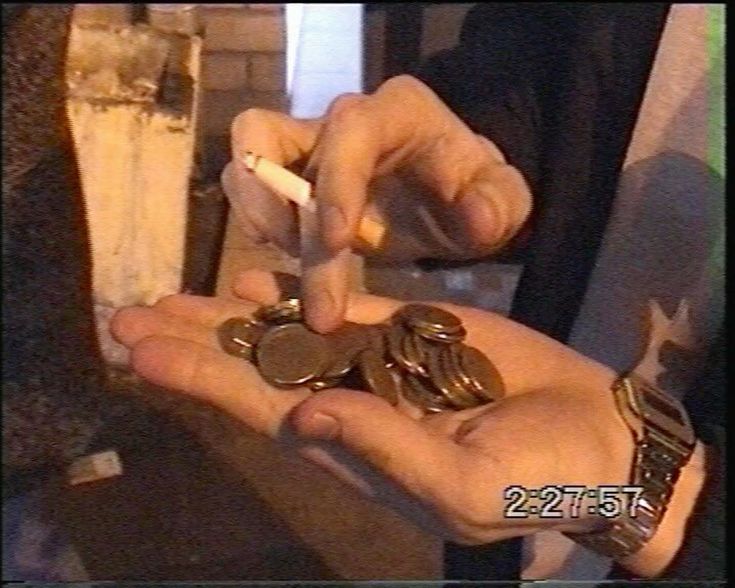 A person holds several coins in one hand while pointing at them with a cigarette in the other. The time “2:27:57” is displayed in the bottom right corner of the dimly lit scene.
