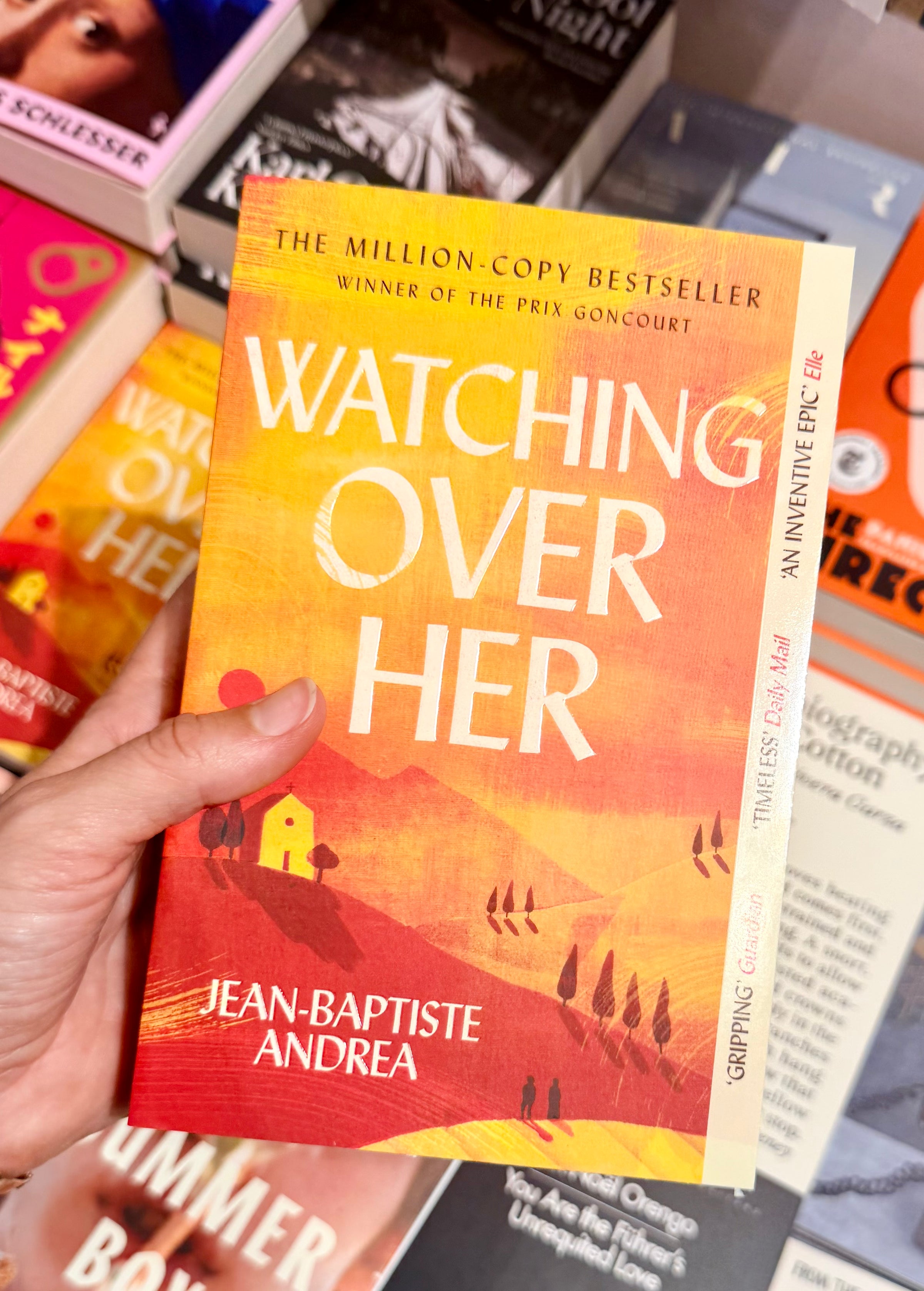 A hand holds the book Watching Over Her by Jean-Baptiste Andrea, featuring a red and yellow cover with a hillside and house, among other books on display.