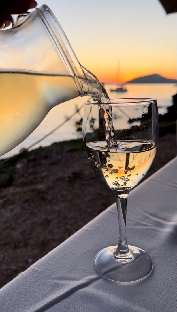 A glass pitcher pours white wine into a wine glass on a table by the water at sunset, with a sailboat and island visible in the background.
